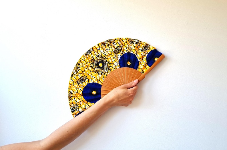May include: A hand fan featuring a vibrant yellow and white geometric pattern with circular blue accents. The fan is constructed with a light brown wooden frame that radiates from the center, supporting the patterned fabric canopy in a semi-circle.