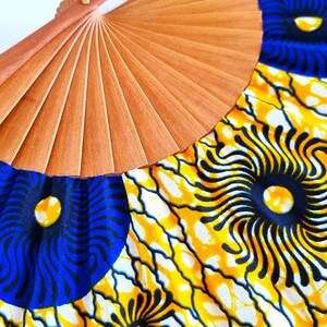 May include: A hand fan featuring a light brown wooden frame with pleated fabric. The fabric displays a vibrant pattern of large blue circular motifs with black radiating lines, set against a bright yellow background with black web-like veins.
