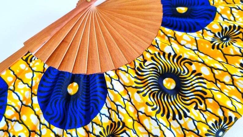 May include: A hand fan featuring a light brown wooden frame with pleated fabric. The fabric displays a vibrant pattern of large blue circular motifs with black radiating lines, set against a bright yellow background with black web-like veins.