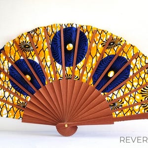 May include: A hand fan featuring a vibrant yellow fabric with a black net-like pattern and dark blue circular motifs. The fan has brown wooden ribs that radiate from a central pivot point. The word REVERSE is printed in the bottom right corner.