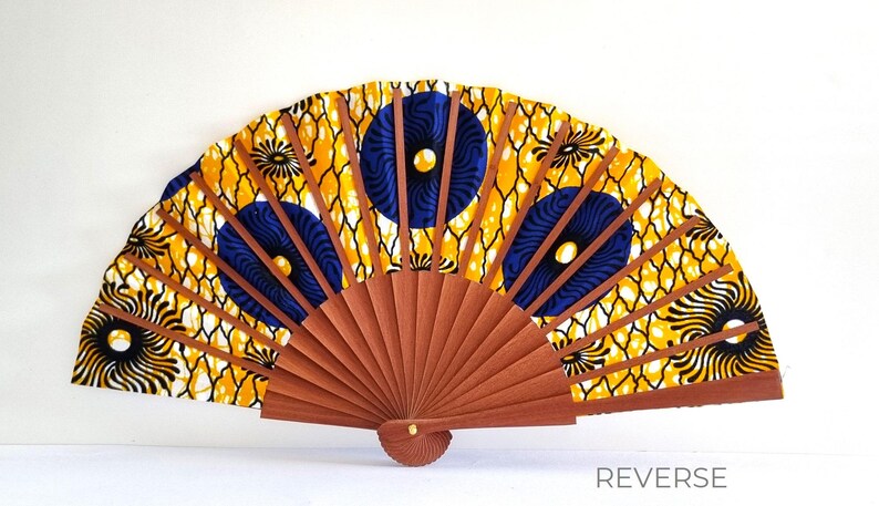 May include: A hand fan featuring a vibrant yellow fabric with a black net-like pattern and dark blue circular motifs. The fan has brown wooden ribs that radiate from a central pivot point. The word REVERSE is printed in the bottom right corner.