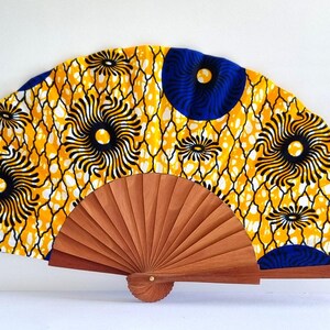 May include: A folding hand fan featuring a vibrant yellow fabric canopy with a repeating pattern of dark blue circular motifs and white net-like lines. The fan is constructed with a light brown wooden frame that radiates from a central pivot point.