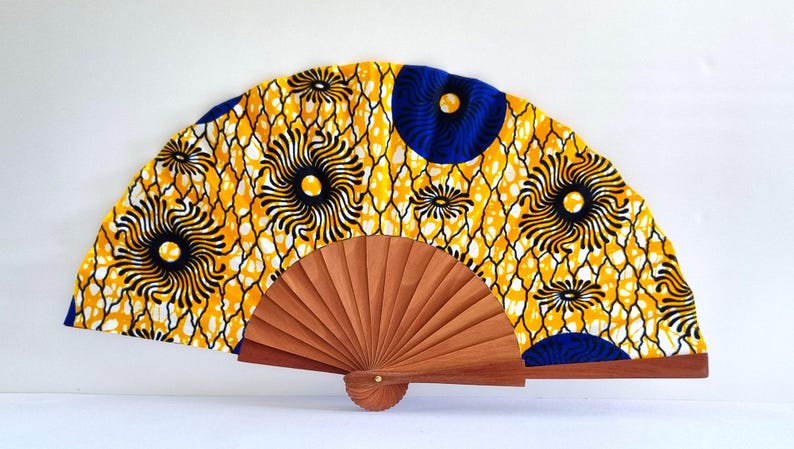May include: A folding hand fan featuring a vibrant yellow fabric canopy with a repeating pattern of dark blue circular motifs and white net-like lines. The fan is constructed with a light brown wooden frame that radiates from a central pivot point.