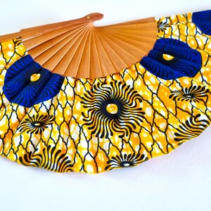 May include: A hand fan featuring a light brown wooden frame and a fabric leaf with a vibrant yellow, black, and blue geometric and floral pattern. The fabric displays a repeating design of blue circular motifs and black radiating lines on yellow.