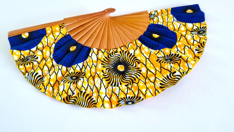 May include: A hand fan featuring a light brown wooden frame and a fabric leaf with a vibrant yellow, black, and blue geometric and floral pattern. The fabric displays a repeating design of blue circular motifs and black radiating lines on yellow.