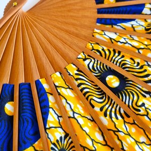 May include: A folding hand fan with light brown wooden ribs and a fabric leaf featuring a vibrant pattern of yellow, black, and blue circular motifs. The fan is partially open, displaying the structural wooden frame and the colorful textile design.