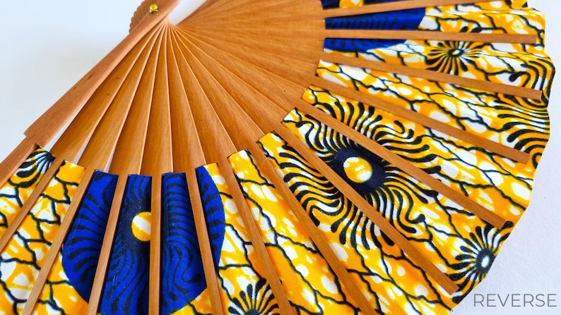 May include: A folding hand fan with light brown wooden ribs and a fabric leaf featuring a vibrant pattern of yellow, black, and blue circular motifs. The fan is partially open, displaying the structural wooden frame and the colorful textile design.