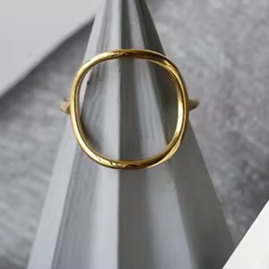 May include: A gold ring with a simple, rounded oval shape. The ring is displayed on a gray cone-shaped stand.