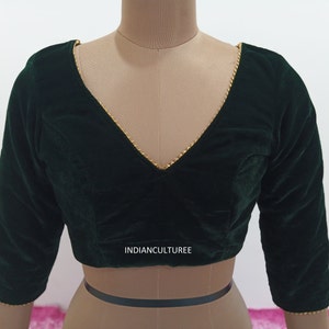 May include: A dark green velvet blouse with a V-neckline and gold trim. The blouse has three-quarter sleeves and a cropped fit. The text "INDIANCULTUREE" is printed on the front of the blouse.