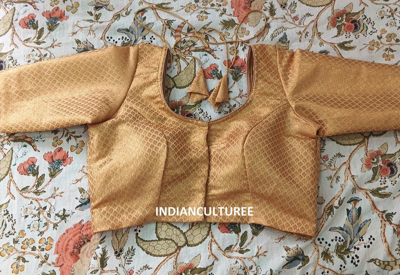 May include: A gold brocade blouse with a square pattern and short sleeves. The blouse has a round neckline and a hook and eye closure in the front. The blouse is decorated with two tassels on the back.