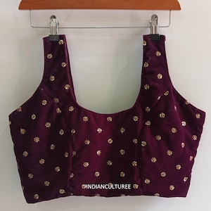 May include: A sleeveless, plum-colored velvet top with gold floral embellishments. The top is hanging on a wooden hanger. The text "INDIANCULTUREE" is visible on the bottom right of the top.