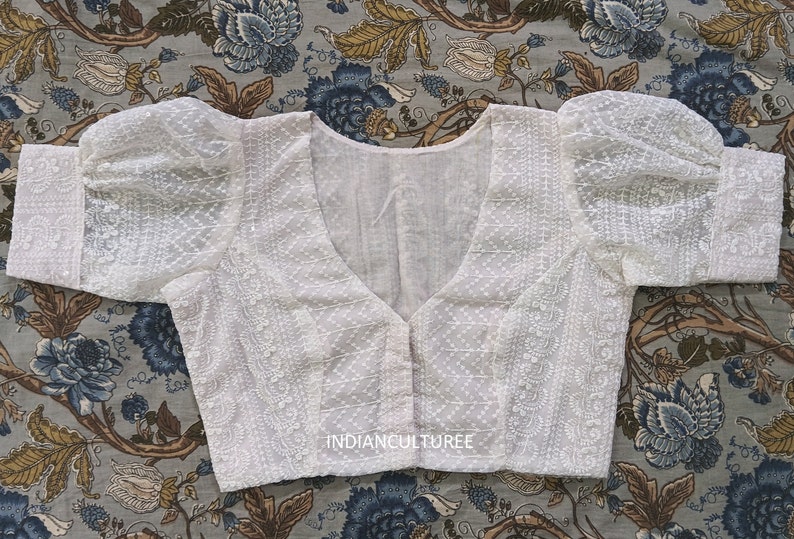 May include: White cotton embroidered blouse with a V-neckline and puffed sleeves. The blouse has a button closure at the front and is decorated with a delicate floral pattern. The text "INDIANCULTUREE" is visible at the bottom of the blouse.