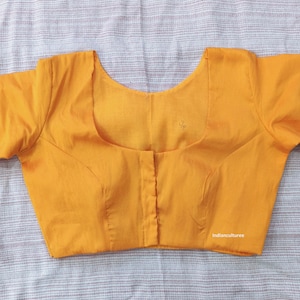 Puede incluir: Blusa de color amarillo mostaza con cuello redondo y mangas cortas. La blusa tiene una silueta entallada con costuras verticales y una botonadura frontal. La tela parece ser un material liso y tejido. El texto "Indianculturee" es visible en la parte inferior derecha.