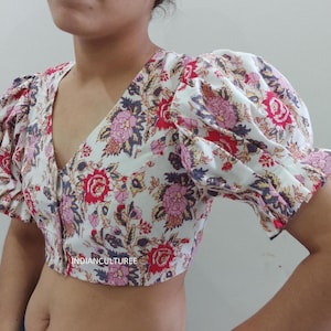 May include: A white crop top with a floral print featuring pink, red, and blue flowers. The top has a V-neckline and short, puffed sleeves. The text "INDIANCULTUREE" is visible on the bottom of the top.