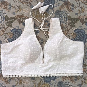 May include: A white, embroidered top with a deep V-neckline and a scalloped hem. The top has a delicate, dotted pattern and adjustable straps. The design is a sleeveless style, perfect for warm weather or layering.