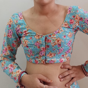 Könnte beinhalten: Eine hellblaue Bluse mit Blumenmuster und Knopfverschluss. Die Bluse hat lange Ärmel und einen runden Ausschnitt.
