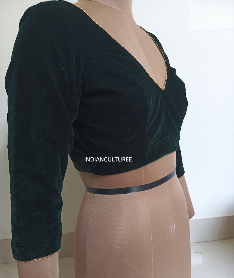 May include: A dark green velvet blouse with gold trim around the neckline and sleeves. The blouse has a V-neck and three-quarter sleeves. The text "INDIANCULTUREE" is visible on the blouse.
