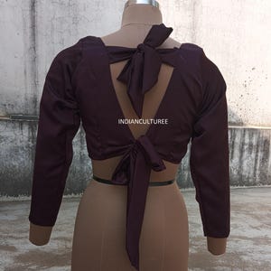 Könnte beinhalten: Eine dunkelviolette Satinbluse mit langen Ärmeln und einem tiefen V-Ausschnitt. Die Rückseite der Bluse hat eine große Schleife, die in der Taille gebunden werden kann. Der Text "INDIANCULTUREE" ist auf der Rückseite der Bluse aufgedruckt.