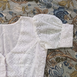 May include: White cotton blouse with a delicate embroidered design and puffed sleeves. The blouse has a V-neckline and a button closure.