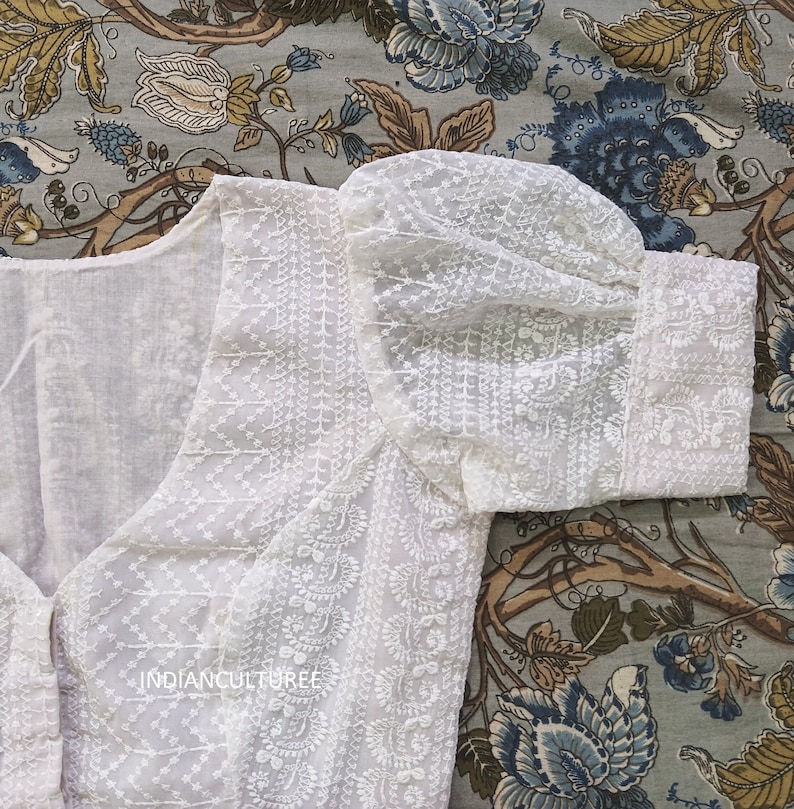 May include: White cotton blouse with a delicate embroidered design and puffed sleeves. The blouse has a V-neckline and a button closure.