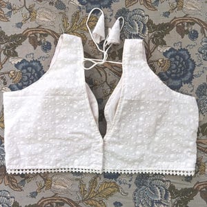 May include: White, sleeveless top with intricate embroidered detailing. The top features a V-neck design and a scalloped trim along the bottom edge. A delicate tie closure is visible at the back. The garment is made of a lightweight fabric.