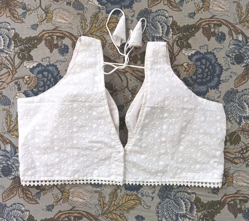 May include: White, sleeveless top with intricate embroidered detailing. The top features a V-neck design and a scalloped trim along the bottom edge. A delicate tie closure is visible at the back. The garment is made of a lightweight fabric.
