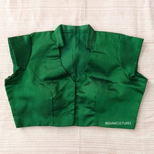 May include: A green, sleeveless blouse with a mandarin collar and a V-neckline. The blouse has a button closure at the front and is made of a smooth, silky fabric. The text "INDIANCULTUREE" is visible at the bottom right corner of the image.
