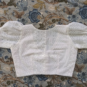 May include: White cotton blouse with a round neckline and puffed sleeves. The blouse has a delicate embroidered pattern and the text "INDIANCULTUREE" is printed on the front.