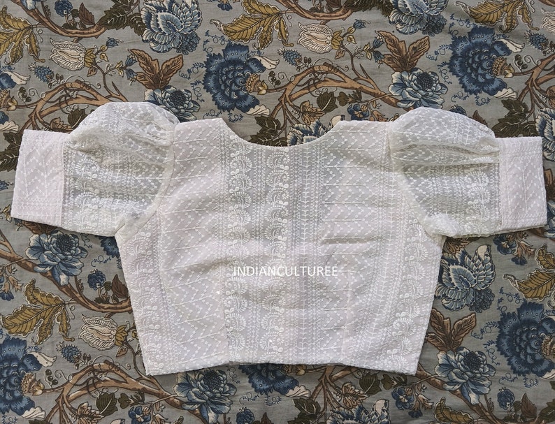 May include: White cotton blouse with a round neckline and puffed sleeves. The blouse has a delicate embroidered pattern and the text "INDIANCULTUREE" is printed on the front.