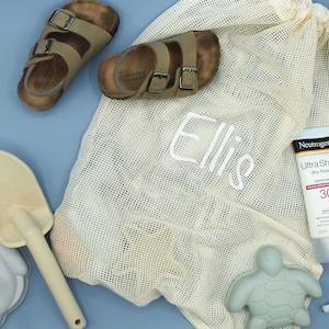 Könnte beinhalten: Eine weiße Netztasche mit dem Namen "Ellie" bestickt. Die Tasche enthält ein Paar braune Sandalen, eine Flasche Sonnencreme, ein grünes Schildkrötenspielzeug und eine Holzschaufel.