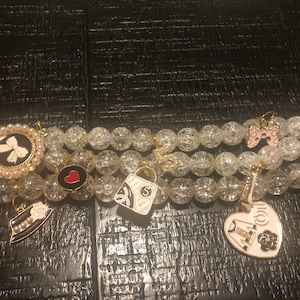 May include: A clear crystal beaded Apple Watch band with multiple charms. The charms include a black and white bow, a red heart, a black and white handbag, a pink bow, and a white heart with a black and white dress and flower.