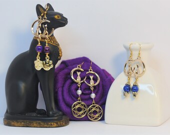 Puede incluir: Tres pares de pendientes de oro colgantes con diferentes encantos. Los pendientes de la izquierda tienen cuentas moradas y encantos de huella de pata. Los pendientes del medio tienen una perla blanca y un encanto dorado con un diseño circular. Los pendientes de la derecha tienen cuentas azules y un encanto dorado con una silueta de gato.