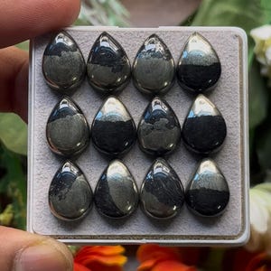 May include: A collection of twelve polished, teardrop-shaped hematite cabochons arranged in a three-by-four grid within a white display box. Each stone features a smooth, metallic grey surface with distinct dark, matte-textured bands.