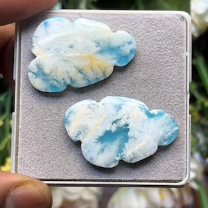 Può includere: Due pietre preziose levigate a forma di nuvola con un motivo marmorizzato nei colori azzurro, bianco e giallo pallido. Le pietre hanno una superficie liscia e lucida e sono disposte fianco a fianco su un vassoio quadrato grigio rivestito in feltro.
