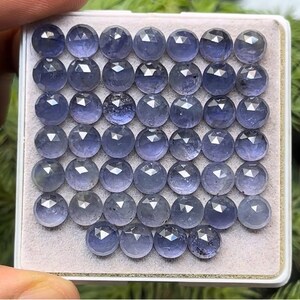 Puede incluir: Una colección de gemas redondas y facetadas en tonos azules y morados. Las gemas están dispuestas en una cuadrícula dentro de una bandeja blanca. Cada piedra tiene múltiples facetas, reflejando la luz. La presentación general es ordenada y organizada.