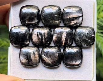 Pode incluir: Uma coleção de dez pedras preciosas pretas polidas, de corte quadrado. Cada pedra tem um brilho metálico reflexivo com padrões internos sutis. As pedras preciosas estão dispostas em uma grade dentro de uma bandeja de cor clara.