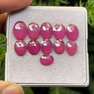 May include: A collection of ten oval-cut, faceted, pink gemstones arranged in a rectangular tray. The gemstones have a polished, reflective surface, and are displayed against a neutral background. The gemstones are a vibrant shade of pink.
