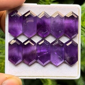 May include: Ten faceted amethyst gemstones in a white tray. The gemstones are a deep purple color with a hexagonal shape. The facets catch the light, creating a sparkling effect. The gemstones are arranged in two rows of five.