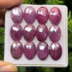 Pode incluir: Uma coleção de doze pedras preciosas de rubi ovais e facetadas. Os rubis são de uma cor vermelho-rosada profunda e estão dispostos numa bandeja branca retangular. As facetas criam um efeito cintilante, realçando a clareza e o brilho das gemas.