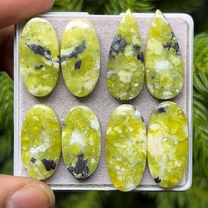 May include: Eight polished gemstones in shades of green and yellow, with dark gray and black inclusions. The stones are oval and teardrop shapes, displayed in a white tray. The gemstones are likely for jewelry making or collecting.