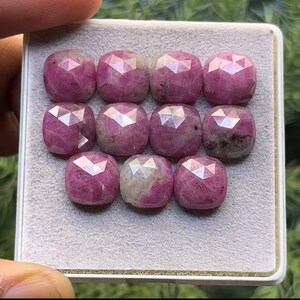 May include: A collection of 10 faceted pink and white gemstones displayed in a white box. The gemstones are all cut in a similar shape and have a polished finish.