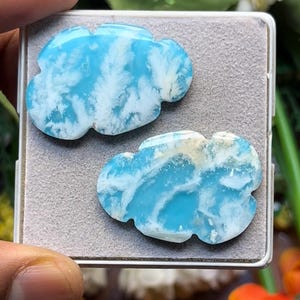 Op de afbeelding: Twee gepolijste, wolkvormige stukken blauw-witte Larimar edelsteen. De stenen hebben een glad, glanzend oppervlak met ingewikkelde, wervelende patronen van hemelsblauw en ondoorzichtig wit, samen gepresenteerd in een klein, vierkant grijs doosje.
