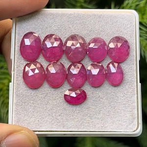 Op de afbeelding: Een verzameling van elf gefacetteerde, ovaalvormige robijnen, gepresenteerd in een witte bak met een grijze binnenkant. De edelstenen vertonen een levendige rozerode tint en zijn in twee rijen gerangschikt, met één robijn onderaan.