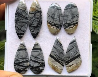 May include: A collection of eight polished gemstones in various shapes: oval, teardrop, and triangular. The stones are primarily gray with intricate black veining and tan accents. They are displayed in a white tray.