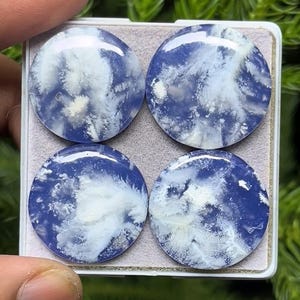 May include: Four round, polished gemstones with a blue and white cloud-like pattern. The stones are set in a rectangular tray, showcasing their unique color variations. The gemstones have a glossy finish.