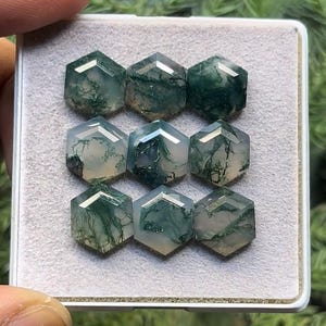 May include: A collection of eight green moss agate cabochons, each cut into a hexagonal shape. The stones are translucent and have a variety of green and white patterns.