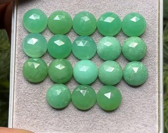 May include: A collection of round, faceted green gemstones displayed in a gray tray. The gemstones vary in shades of green, from light to deep, and have a polished, reflective surface. The tray is set against a blurred background.