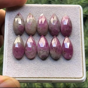 May include: Ten pink pear-shaped gemstones with a faceted cut. The gemstones are arranged in two rows of five, and are displayed in a white box.