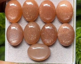 May include: Eight oval-shaped, peach-colored gemstones with a polished, reflective surface are displayed in a white tray. The gemstones have a subtle shimmer and varying degrees of translucence. One additional gemstone is positioned below the others.
