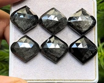 Pode incluir: Seis pedras preciosas pretas, facetadas em forma de diamante, são exibidas numa bandeja branca. As pedras têm uma aparência reflexiva e metálica, com padrões internos visíveis. A bandeja é segurada por uma mão, com folhagem verde ao fundo. Cada gema mede cerca de 1,5 cm.